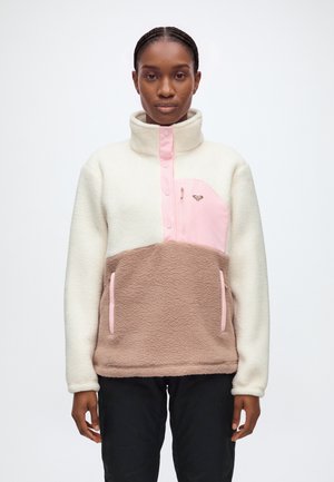 Une femme porte un pull polaire à blocs de couleur crème, rose et taupe, avec un col montant et une poche à l’avant, associé à un pantalon noir.
