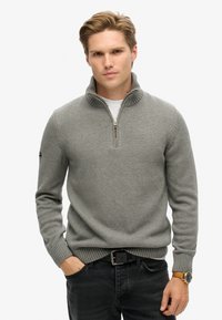 Grauer Strickpullover mit hohem Kragen und einem Viertelreißverschluss. Mit gerippten Bündchen und Saum, über einem weißen T-Shirt getragen. Schwarze Jeans.