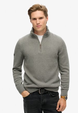 Pull en maille grise avec un col montant et une fermeture éclair quart. Manches et ourlet côtelés, porté par-dessus un t-shirt blanc. Jean noir.