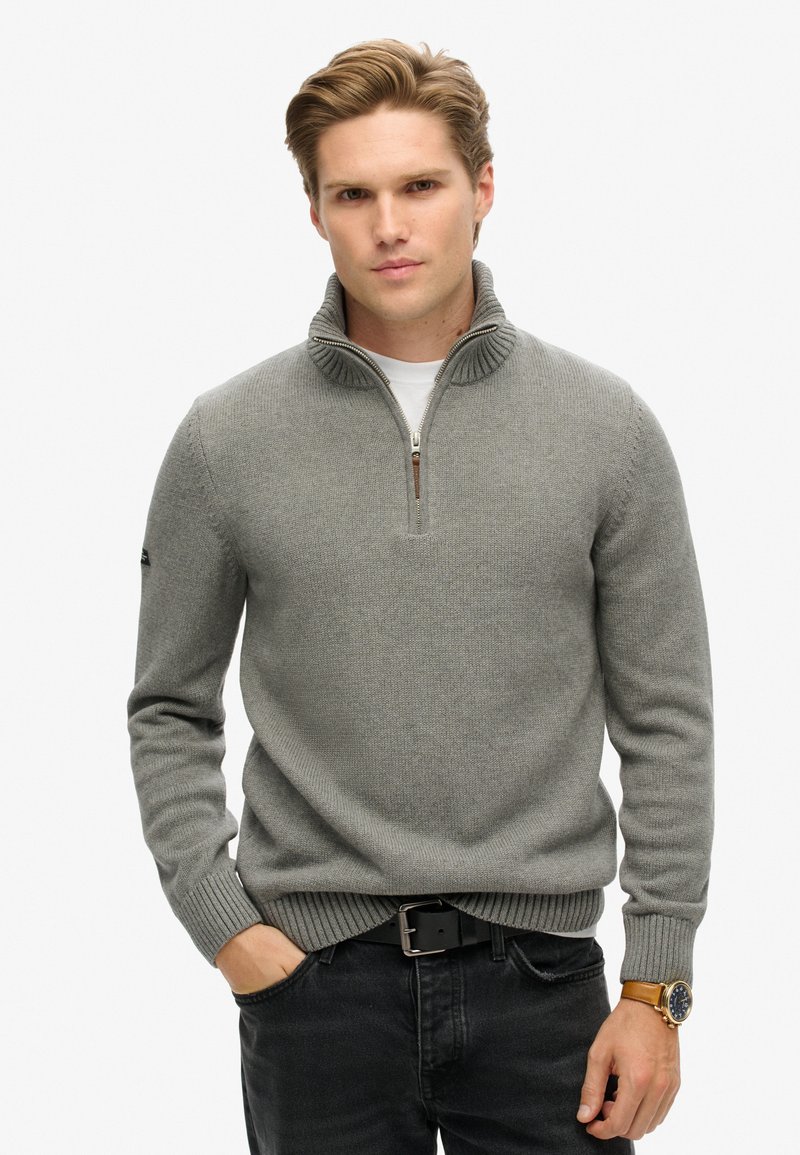 Grauer Strickpullover mit hohem Kragen und einem Viertelreißverschluss. Mit gerippten Bündchen und Saum, über einem weißen T-Shirt getragen. Schwarze Jeans.