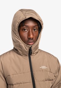 Tan puffer jacket with a high collar and hood, featuring a black zipper and logo on the chest. Soft texture and quilted design.
