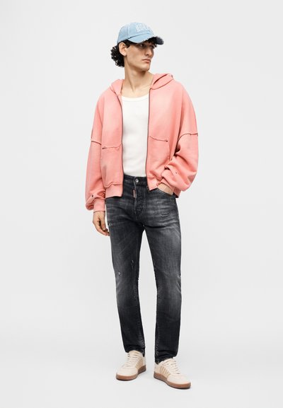 Φούτερ σε ροζ ανοιχτό χρώμα σε υπερμεγέθη γραμμή με τσέπες μπροστά, φορεμένο πάνω από λευκό αμάνικο τοπ. Σκούρο τζιν με ξεθωριασμένη όψη και ανοιχτά μπεζ αθλητικά παπούτσια. Μπλε καπέλο.