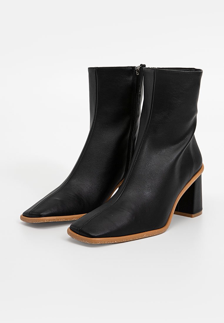 Schwarze Leder-Stiefeletten mit spitzer Zehenform, seitlichem Reißverschluss und blockartigem Absatz. Auffällige glatte Textur und braune Akzentsohle.