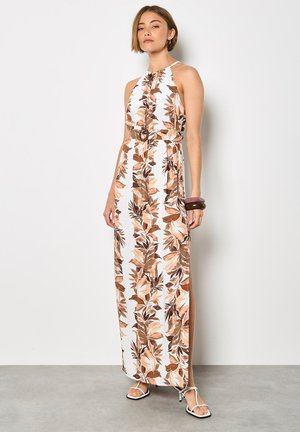 Kobieta w białej sukience maxi bez rękawów z brązowym kwiatowym nadrukiem, z paskiem w talii i białymi sandałkami na obcasie, stoi przy gładkiej ścianie.