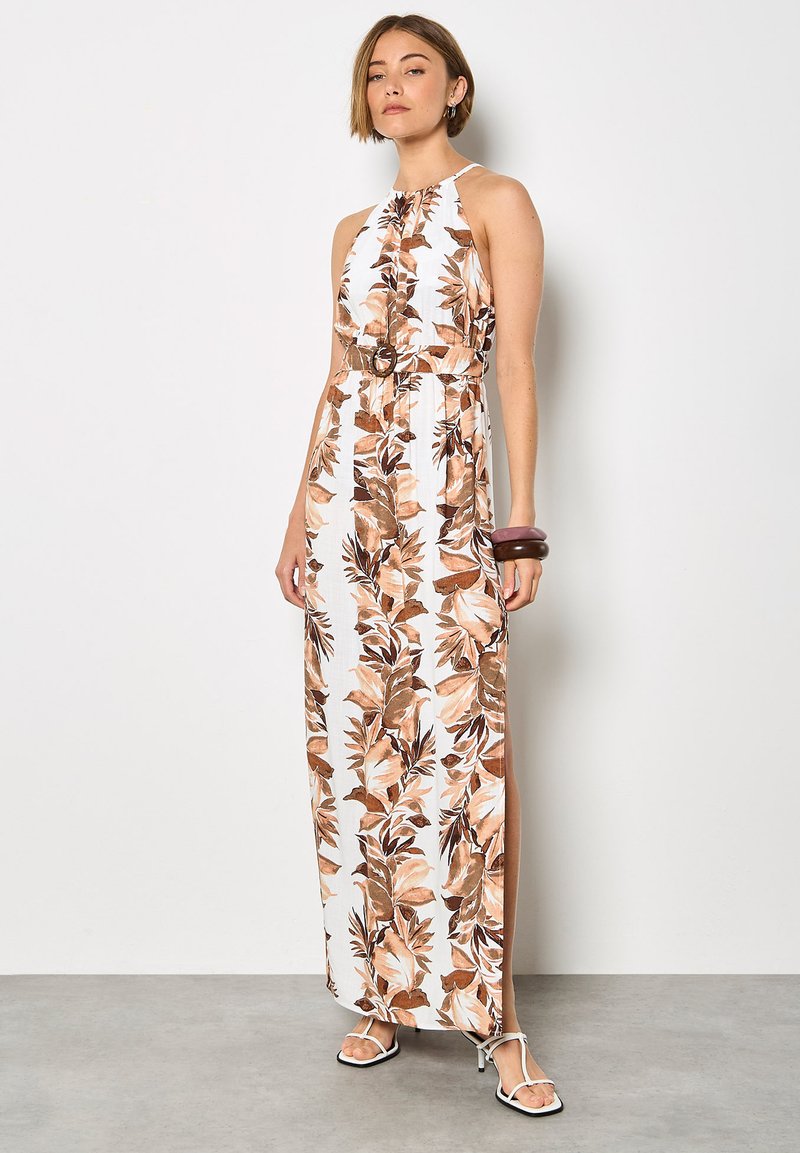 Femme portant une robe longue blanche sans manches avec un imprimé floral marron, taille ceinturée, et des talons blancs à brides, debout contre un mur uni.
