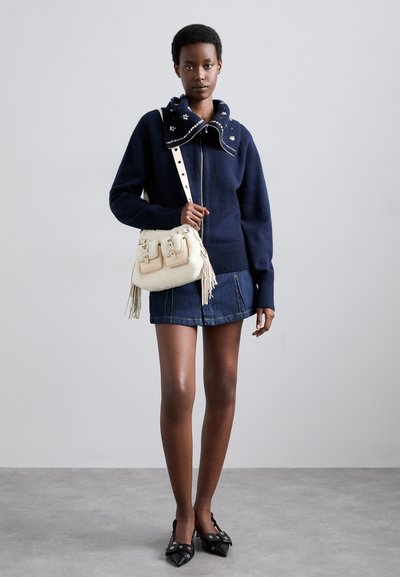 Maglione blu navy con zip e colletto decorato, gonna di jeans, borsa bianca con frange e fibbie, e décolleté nere con punta appuntita.
