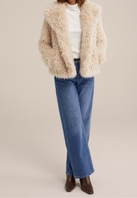 Faux fur-beige jacka med sjalkrage, bärs över en vit topp, tillsammans med ljusblå vida jeans och bruna ankelskapsstövlar.