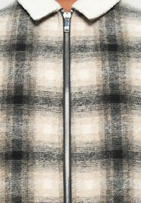 Jachetă cu fermoar, cu model tartan, având un guler cald și pufos. Culorile includ gri, negru și bej moale. Fabricată dintr-un material texturat.