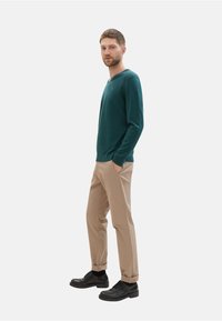 Pull en maille verte, en V, pantalons beiges, chaussures noires, pose debout, mains dans les poches. Design minimaliste sur fond blanc.