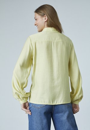 Gul langærmet skjorte lavet af letvægtsstof med rygpanel og elastiske manchetter, parret med højtaljede blå jeans.