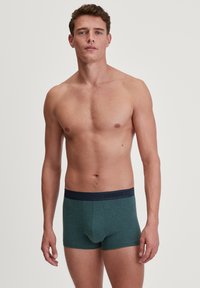 Herenboxers in donkergroene gemêleerde stof, met een aansluitend ontwerp, voorzien van een effen marineblauwe tailleband met subtiele lettering van de merknaam "CALIDA".