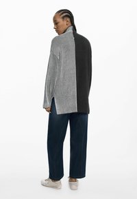 Pull en maille avec un col montant, présentant des sections rayées noir et gris. Associé à un pantalon en denim ample et des baskets de couleur claire.