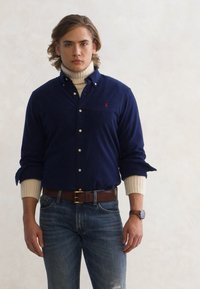 Jovem vestido com uma camisa abotoada azul-marinho sobre um suéter creme, jeans azuis, cinto castanho e relógio de pulso, de pé contra um fundo liso.