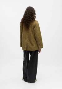 Olijfgroene, geweven oversized blazer met een rugventilatie en knoopmanchetten, gecombineerd met een brede, donkere spijkerbroek en neutrale canvas schoenen.