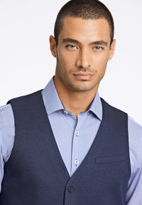 Marineblau Wolleweste, die über einem hellblau-weiß gestreiften Hemd getragen wird, mit einer Tasche und einem einzigen Knopfverschluss. Anspruchsvolle Designdetails.