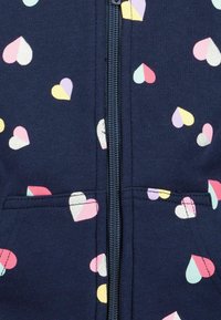 Navy zip-up hoodie featuring a repeated pattern of colorful, varying-sized hearts, including metallic accents, with front pockets.