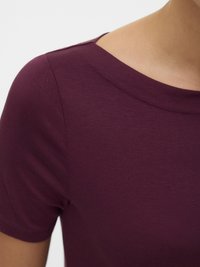 Krupan plan osobe koja nosi majicu kratkih rukava boje tamnocrvene, prikazujući izrez i područje ramena na svijetloj pozadini.