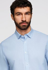 Homme aux cheveux bouclés foncés et barbe portant une chemise à boutons bleu clair, face à l'avant sur un fond clair uni.