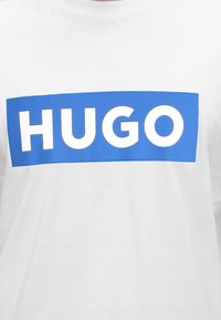 Chemise blanche avec un logo rectangulaire bleu affichant le mot "HUGO" en lettres blanches en gras sur la poitrine.