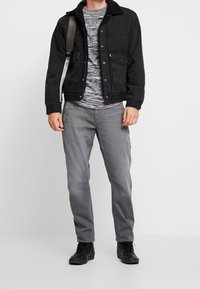 Veste en denim noire avec un col en fourrure, pull à rayures grises, pantalon gris et baskets noires, avec une bandoulière sur la veste.