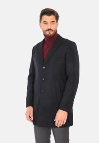 Cappotto di lana blu navy con design a un petto, tre bottoni, due tasche frontali e rever a taglio. Texture liscia con vestibilità su misura.