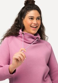 Felpa rosa con collo alto, dotata di un cordino con fermagli marroni. Texture liscia e vestibilità comoda con polsini a coste.