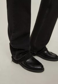 Jeans di denim neri con orli sfrangiati e mocassini in pelle nera con una parte superiore texturizzata e suole piatte, visti dalla caviglia in giù.