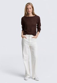 Barna kötött pulóver széles nyakkivágással, magas derekú fehér farmerrel párosítva. A modell fehér tornacipőt visel, és egyenesen áll, kezei a zsebekben.