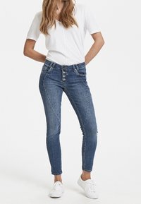 Tajetas ajustadas de mezclilla azul medio con un cierre de cinco botones, que presentan costuras anguladas y un diseño entallado, combinadas con una camiseta blanca lisa.