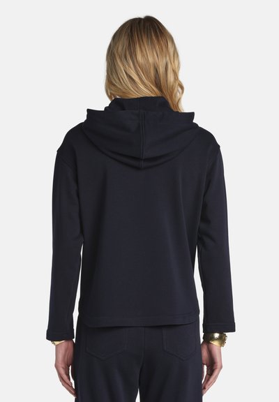 Szőke személy, egyszínű fekete kapucnis pulóvert és hozzáillő fekete nadrágot visel, hátulnézetben, fehér háttér előtt.