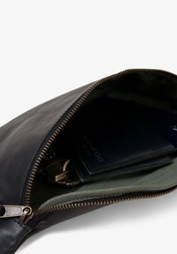 Schwarzes Lederetui mit bronzefarbenem Reißverschluss. Das Innere zeigt ein grünes Futter, Schlüssel und zwei dunkle Geldbörsen. Glatte Textur mit minimalistischem Design.