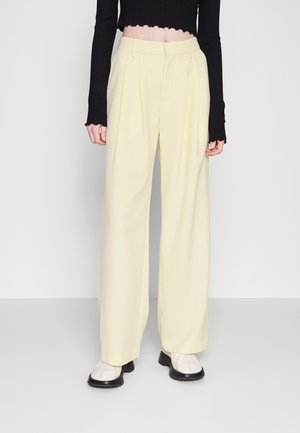 Personne portant un pantalon large crème taille haute, un haut noir à manches longues court, et des chaussures blanches à semelles noires, debout sur un sol gris.