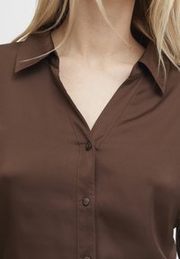 Camicia marrone con bottoni, dotata di un colletto strutturato, presenta una texture liscia, design a scollo a V e due bottoni sul davanti.