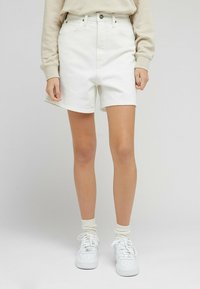 Bílé denimové kraťasy s vysokým pasem, předními kapsami a roztrhaným lemem, v kombinaci s bílými teniskami a světle krémovými ponožkami.