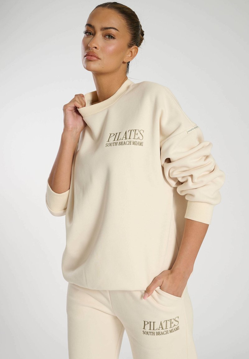 Cremefarbenes übergroßes Sweatshirt mit geripptem Halsausschnitt, das "PILATES SOUTH BEACH MIAMI" in goldener Stickerei zeigt. Weicher, plüschiger Stoff.