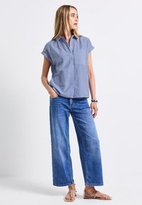 Camicia a maniche corte a righe blu con taschino, abbinata a jeans denim blu a gamba larga e sandali beige con cinturino.