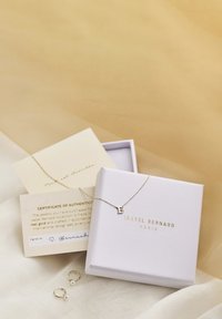 Goldene Halskette mit einem Anhänger in Form des Buchstabens "B", platziert in einer weißen Box mit goldenen Akzenten. Außerdem enthalten sind zwei goldene Creolen und ein Echtheitszertifikat.