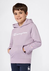Garçon souriant portant un sweat à capuche lilas de la marque Champion, les mains dans la poche avant, et un pantalon navy, se tenant devant un fond blanc uni.