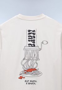 Κρεμ t-shirt με γραφικό μιας μηχανής ζυμαρικών με μαύρα νουντλς και κόκκινη σάλτσα που χύνεται σε ένα πιάτο. Κάτω από το σχέδιο γράφει, «ΦΑΕ ΖΥΜΑΡΙΚΑ. ΤΕΛΟΣ.»