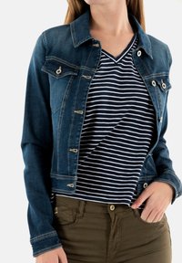 Chaqueta de mezclilla en azul oscuro con una forma ajustada, herrajes plateados y un cuello tipo spread. Llevada sobre una camiseta de rayas en azul marino y blanco con escote en V.