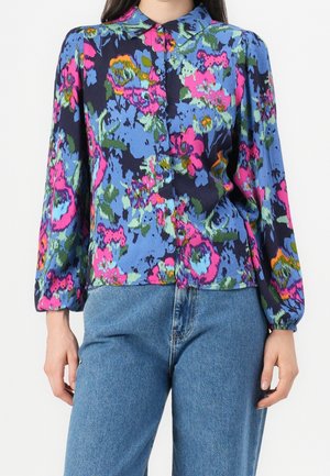 Femme portant un chemisier à manches longues à boutons avec imprimé floral bleu, rose et vert, associé à un jean en denim bleu.