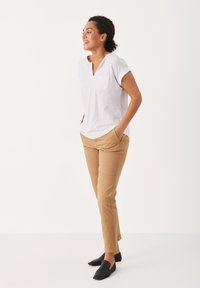 White short-sleeve t-shirt with a front pocket, paired with fitted beige chinos and black slip-on shoes, standing with hands in pockets.