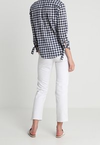 Camisa a cuadros en azul marino y blanco con mangas remangadas, combinada con jeans blancos recortados, que presentan dobladillos desflecados. Llevada con sandalias planas.