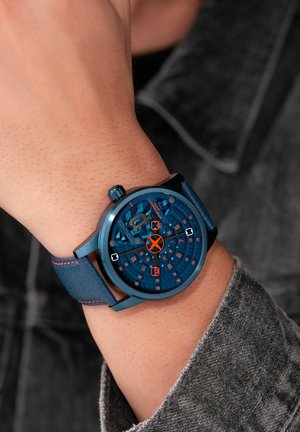 Blaues Analog-Uhr mit rundem Ziffernblatt, strukturiertem blauen Lederarmband, orangenen Akzenten und mehreren Ziffernmarkierungen auf einem detaillierten Zifferblatt.