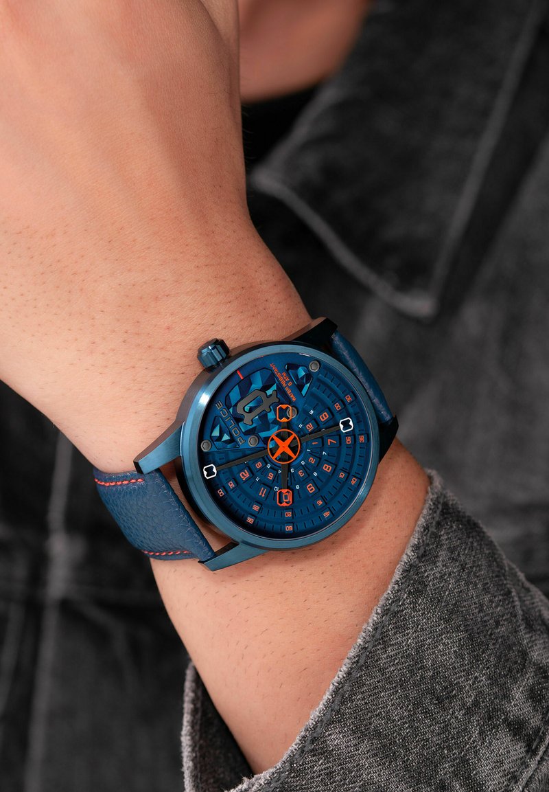 Blauw analoog horloge met een ronde wijzerplaat, een gestructureerde blauwe leren strap, oranje accenten en meerdere cijfermarkeringen op een gedetailleerde wijzerplaat.