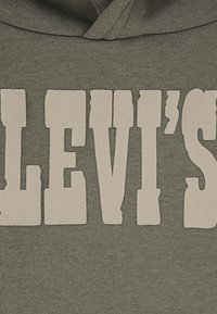 Hoodie vert olive avec un texte blanc bien visible portant "LEVI'S" dans une police audacieuse et rétro. Le tissu semble doux et texturé.