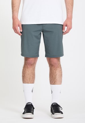 Homme portant un short gris mi-genou, des chaussettes blanches avec un logo noir et des baskets noires basses, debout contre un fond blanc.