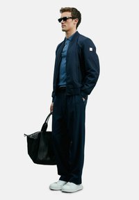Marineblauer Bomberjacke über einem hellblauen Hemd, kombiniert mit dunklen Hosen und weißen Sneakers. Hält eine schwarze Tasche und trägt dunkle Sonnenbrillen.
