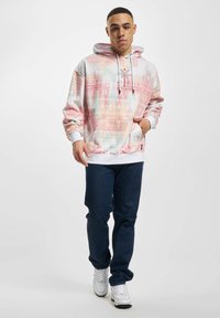 Pullover hoodie with intricate pastel geometric patterns, drawstring hood, front pocket, and white ribbed cuffs and hem. Paired with blue jeans and sneakers.