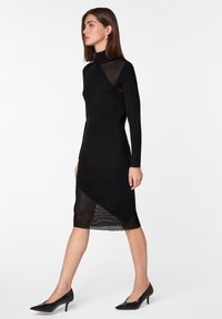 Robe en tricot noire avec un col montant et des manches longues. Le design présente un panneau en maille diagonal à l'ourlet. Associée à des talons noirs.